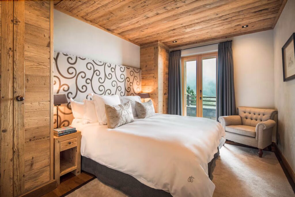 A cozy bedroom with a wooden ceiling and walls, a large bed with white bedding, two bedside tables with lamps, a patterned headboard, a gray armchair by a window, and curtains framing a glass door to a balcony. David Pearson Travel