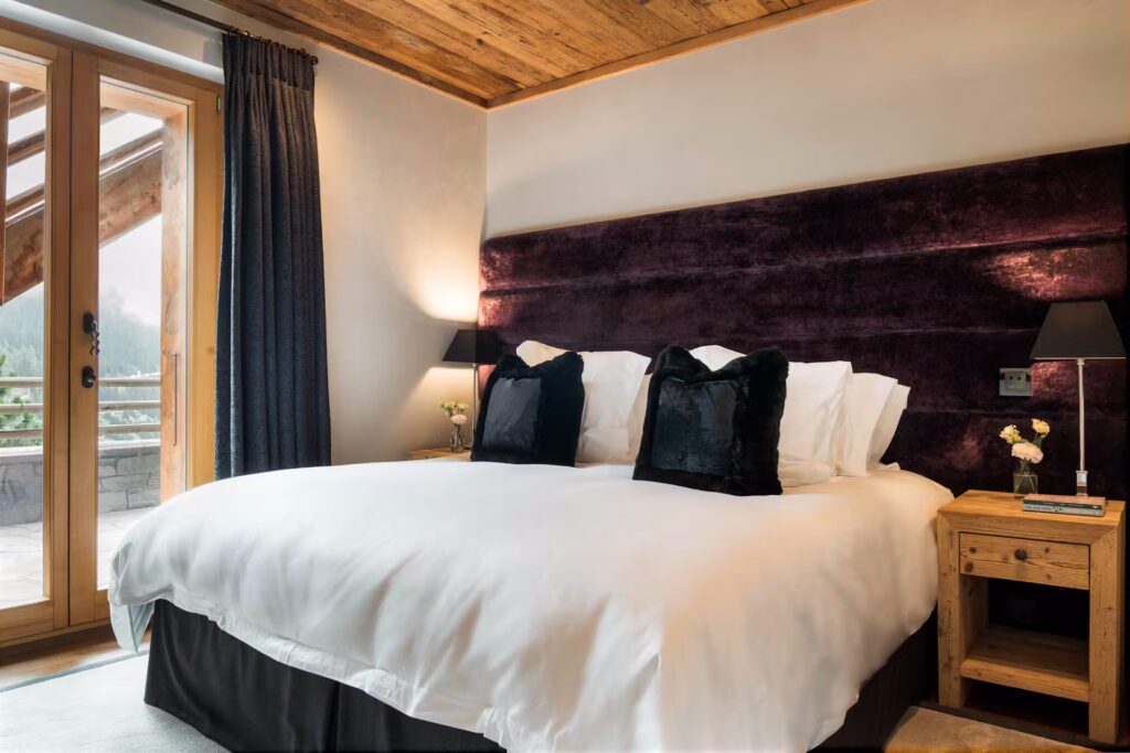 A cozy bedroom with a large bed featuring white linens and black pillows, wooden nightstands with lamps, a dark upholstered headboard, and glass doors leading to a balcony with a view outside. David Pearson Travel