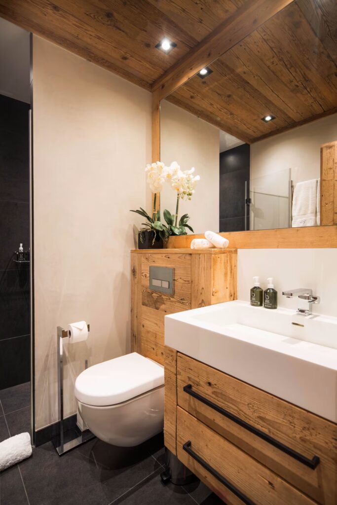 Modern bathroom with wood accents, a wall-mounted toilet, white sink with wooden cabinet, large mirror, black floor tiles, and a potted orchid on top of a wooden shelf next to the toilet. David Pearson Travel