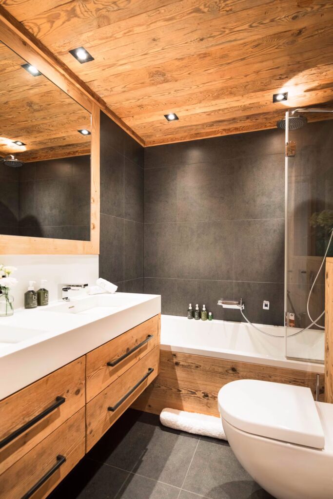 Modern bathroom with wooden ceiling and cabinetry, white sink, wall-mounted toilet, dark tiled walls, bathtub with glass screen, and recessed lighting; decorative bottles and flowers on the vanity. David Pearson Travel