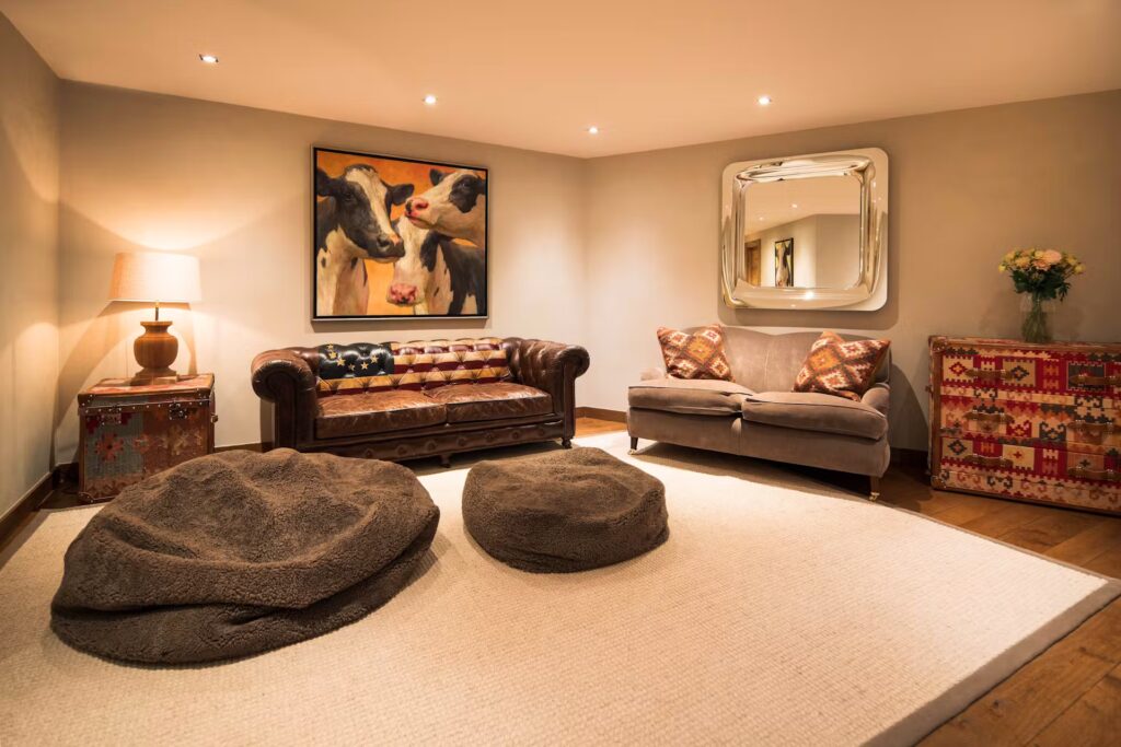 A cozy living room with a leather and a fabric sofa, two large round floor cushions, a rug, a lamp, a colorful side cabinet, a vase of flowers, and artwork of cows on the wall. Warm lighting creates a relaxing atmosphere. David Pearson Travel