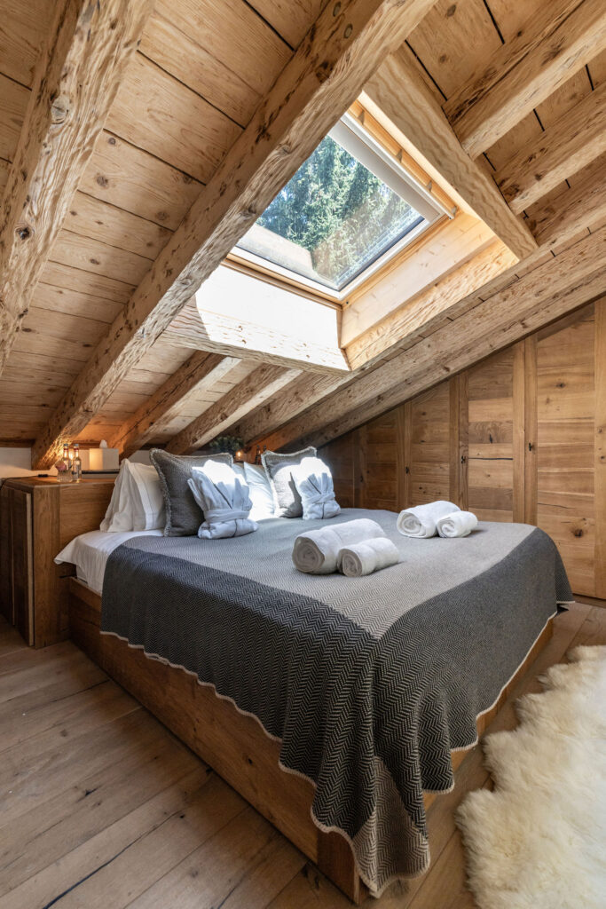 Cozy attic bedroom with a wooden ceiling and walls, a double bed with gray bedding and pillows, rolled towels, a sheepskin rug, and a skylight window letting in natural light. David Pearson Travel