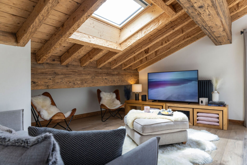 Cozy attic living room with exposed wooden beams, a skylight, a TV on a wooden console, two butterfly chairs with fur throws, an ottoman, and fluffy rugs on light wood flooring. David Pearson Travel