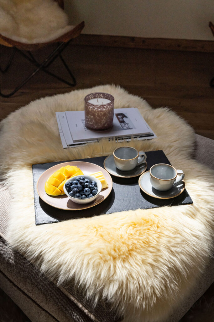 A cozy scene with two cups of coffee, a plate of sliced mango and blueberries, a candle, and magazines on a fluffy cream-colored throw over an ottoman. Sunlight streams onto the inviting setup. David Pearson Travel