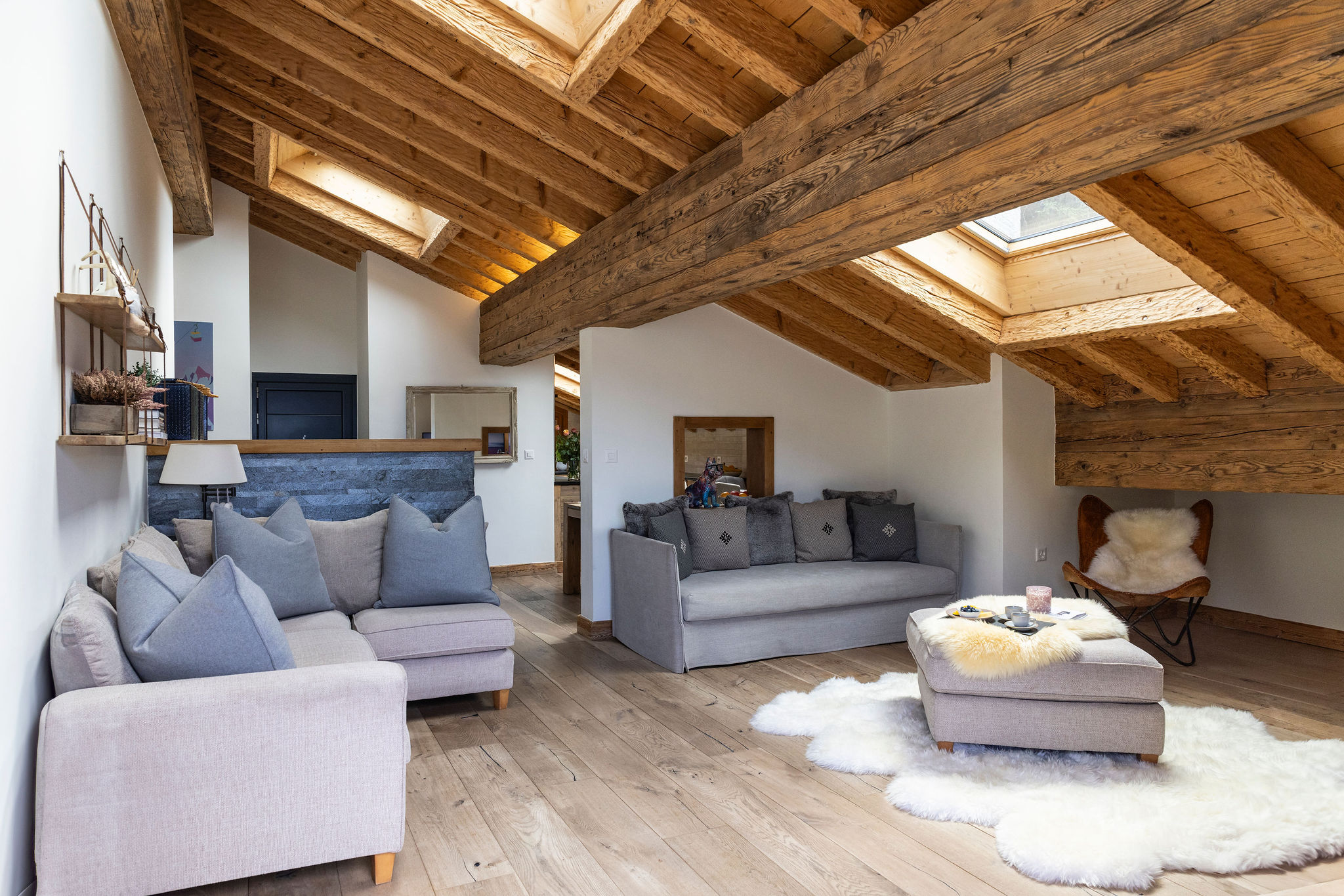 Cozy attic living room with exposed wooden beams, gray sofas, a cushioned ottoman, light wood floors, sheepskin rugs, and a skylight letting in natural light. Shelves and decorative items add a warm, inviting feel. David Pearson Travel