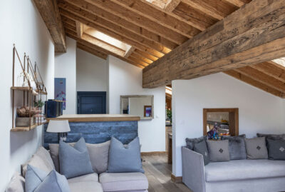 A cozy loft living room with wooden beams, light wood floors, a skylight, two gray sofas with blue and gray cushions, and a small open kitchen area in the background. David Pearson Travel