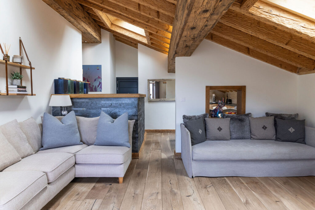 A cozy living room with wooden floors and a vaulted ceiling with exposed beams, featuring two sofas with gray and beige cushions, a small shelf with decor, and a partial stone accent wall. Natural light streams in from skylights. David Pearson Travel