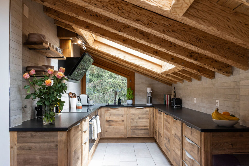 Modern rustic kitchen with wooden cabinets and exposed beams under a sloped ceiling, large window with forest view, black countertops, flowers in a vase, and a bowl of fruit on the counter. David Pearson Travel