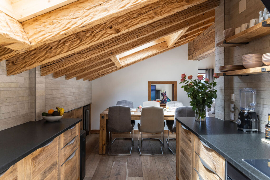 A modern rustic kitchen with exposed wooden beams, open shelving, dark countertops, and a vase of roses. In the background, a dining table with gray chairs sits under a skylight in a bright, cozy space. David Pearson Travel