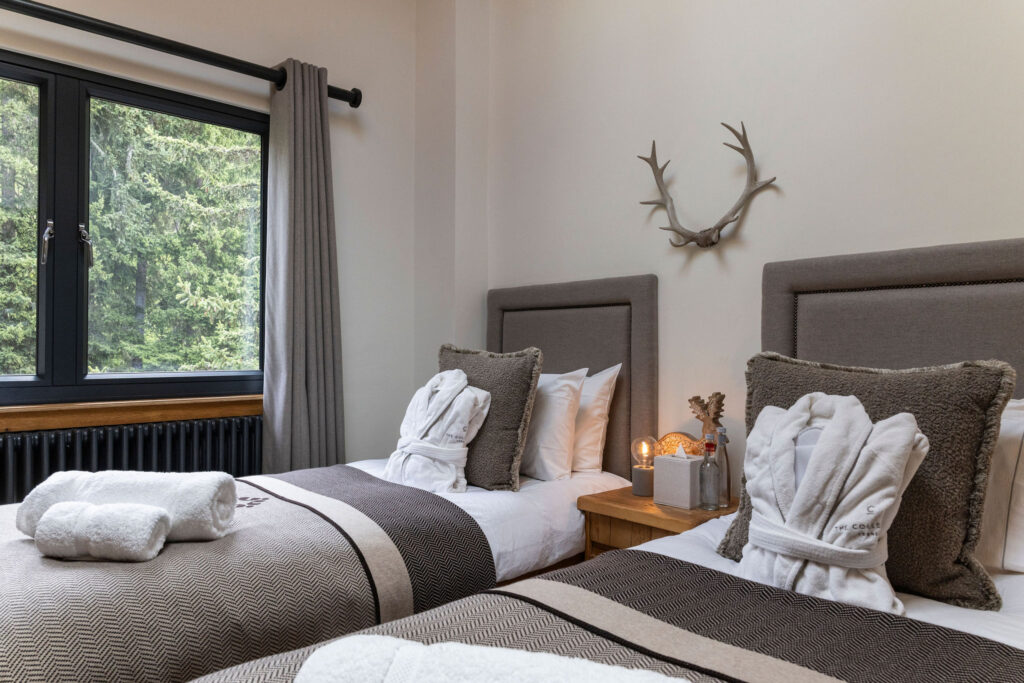 A cozy bedroom with two twin beds, gray headboards, neutral bedding, bathrobes, and towel sets. A wooden nightstand holds decor and candles. A window shows green trees, and antlers hang on the wall above the beds. David Pearson Travel