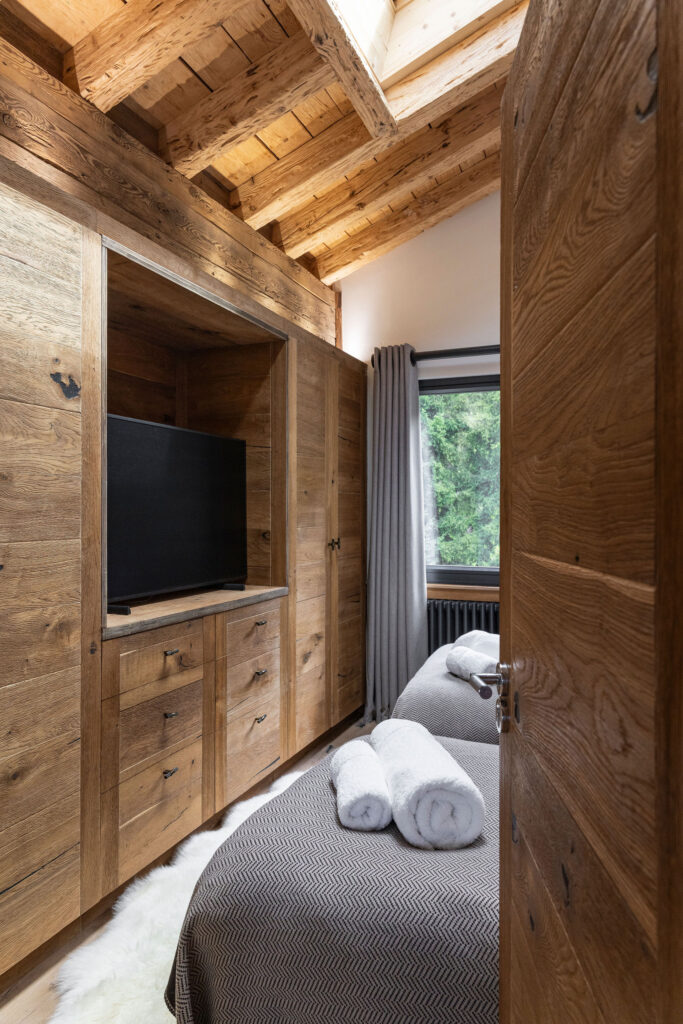 A cozy bedroom with rustic wooden walls and ceiling, featuring a bed with gray bedding and two rolled towels, a built-in wooden wardrobe, a TV, and a window with gray curtains overlooking greenery. David Pearson Travel