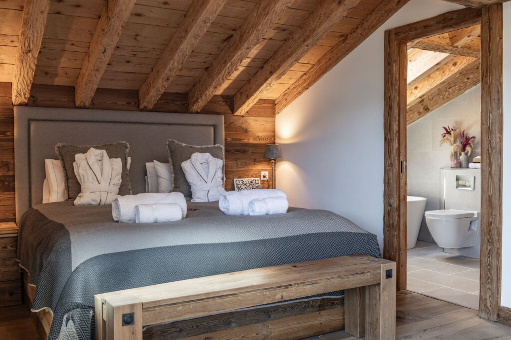 A cozy attic bedroom with exposed wooden beams, a large bed with gray bedding, two bathrobes, and towels. An open door reveals a modern bathroom with a toilet and vase of flowers. Natural light brightens the space. David Pearson Travel