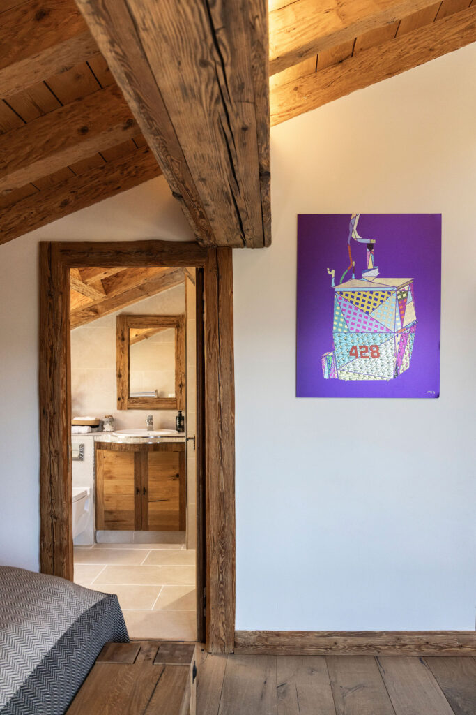 A rustic bedroom with exposed wooden beams and walls, features a modern colorful artwork on a white wall and an open doorway leading to a bathroom with wooden cabinetry and a large mirror. David Pearson Travel