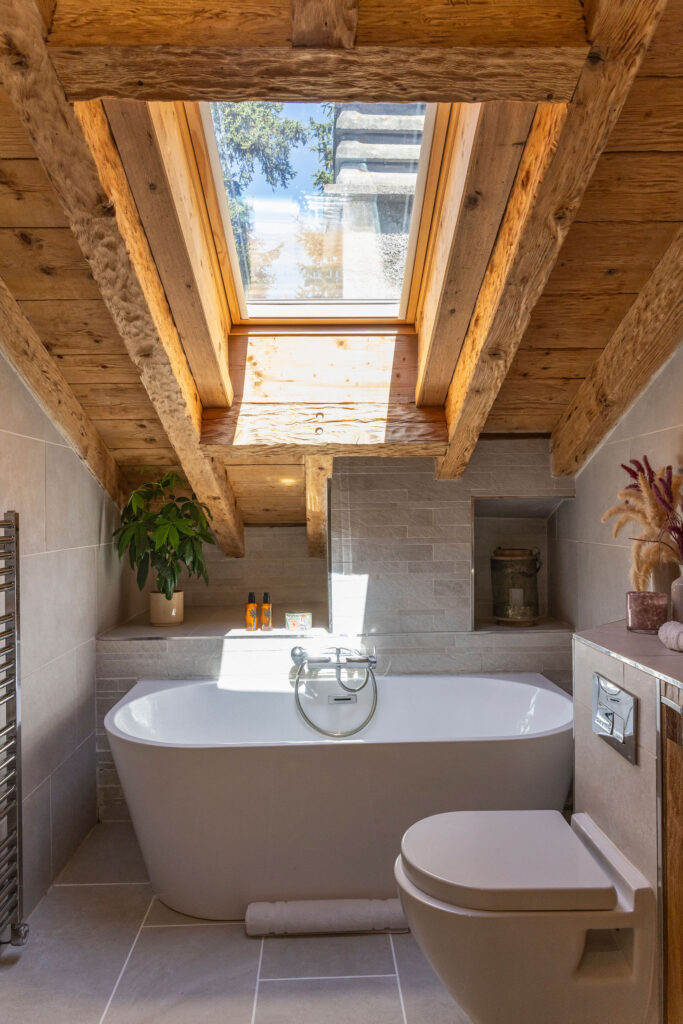 A cozy bathroom with a freestanding bathtub under a skylight window, wooden ceiling beams, light gray tiles, a green potted plant, and neatly arranged toiletries on a shelf. David Pearson Travel