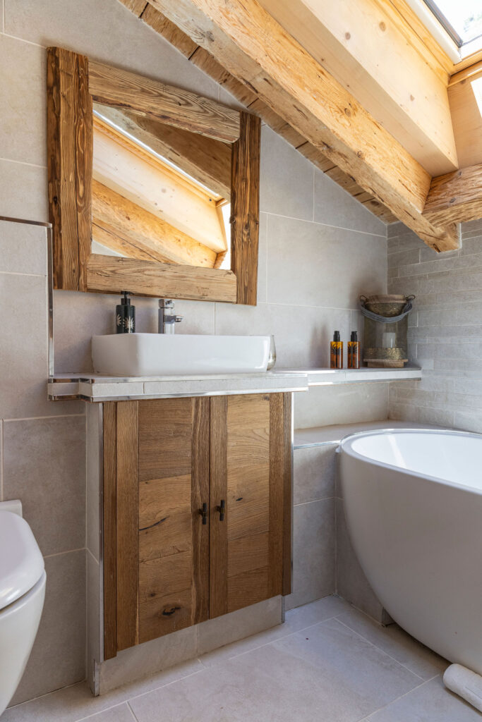 A rustic bathroom with a wooden vanity, vessel sink, rectangular mirror, freestanding bathtub, and large skylight window letting in natural light. Exposed wooden beams and neutral tiles complete the cozy design. David Pearson Travel