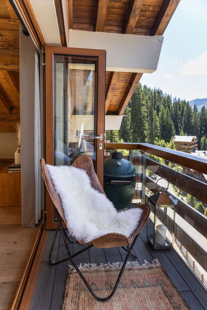 A cozy balcony with a leather chair draped in a white fur throw, a small rug, a lantern, and a green ceramic grill, overlooking a scenic view of trees, houses, and mountains under a clear sky. David Pearson Travel