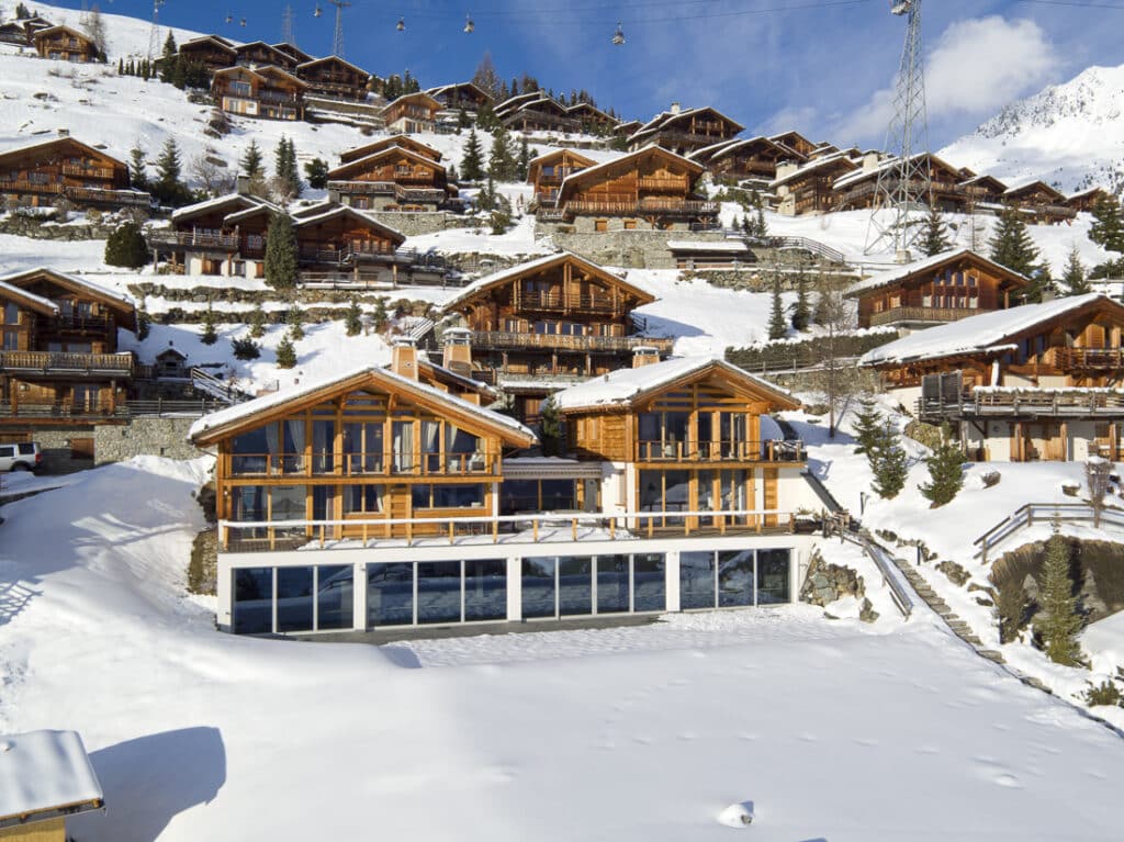 A cluster of wooden alpine chalets sits on a snowy hillside under a blue sky, with snow-covered rooftops and pine trees scattered around, evoking a peaceful winter village scene. David Pearson Travel