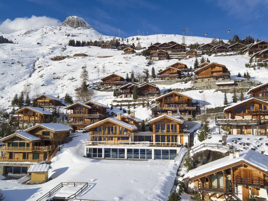Wooden chalets dot a snowy mountain slope under a clear blue sky, with a rocky peak in the background and ski lifts visible above the rooftops. David Pearson Travel