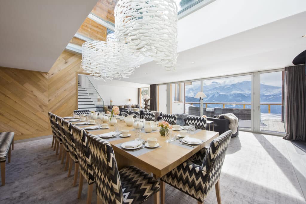 A modern dining room with a long wooden table set for twelve, zigzag-patterned chairs, large white pendant lights, and floor-to-ceiling windows revealing a snowy mountain view. David Pearson Travel