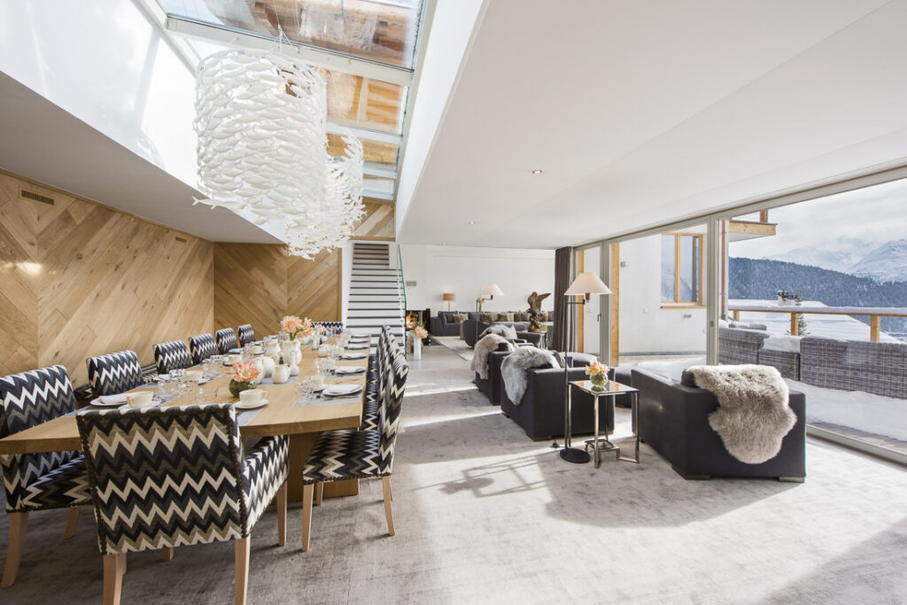 A modern, sunlit open-plan living and dining room with large windows, wooden accents, a zigzag-patterned dining set, cozy sofas with fur throws, and views of a snowy mountain landscape outside. David Pearson Travel