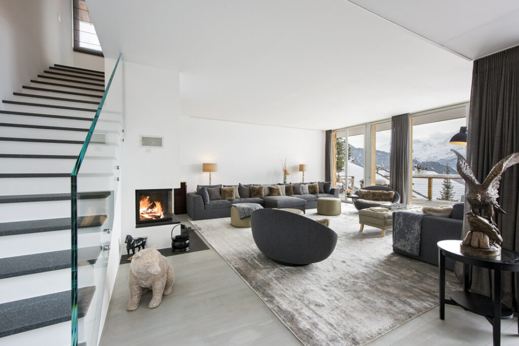 Modern living room with large windows, gray sofas around a fireplace, a round chair, sculpted dog and eagle figures, and a staircase with a glass railing. Mountain scenery is visible through the windows. David Pearson Travel