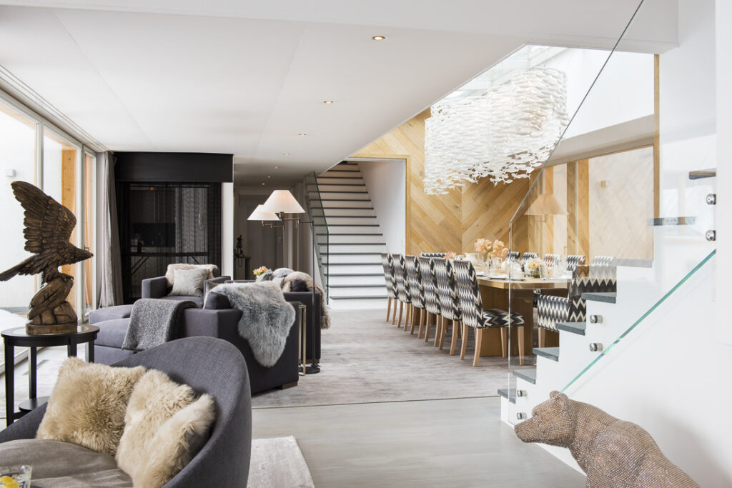 Modern open-plan living and dining area with gray sofas, fur throws, a wooden eagle sculpture, a long dining table with patterned chairs, and a sculptural white chandelier. A staircase with glass railing leads upstairs. David Pearson Travel