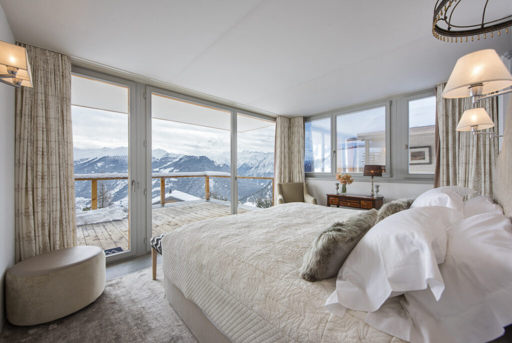 A cozy bedroom with a large bed, soft bedding, and neutral decor features floor-to-ceiling windows and a balcony overlooking snowy mountains and a scenic outdoor view. David Pearson Travel