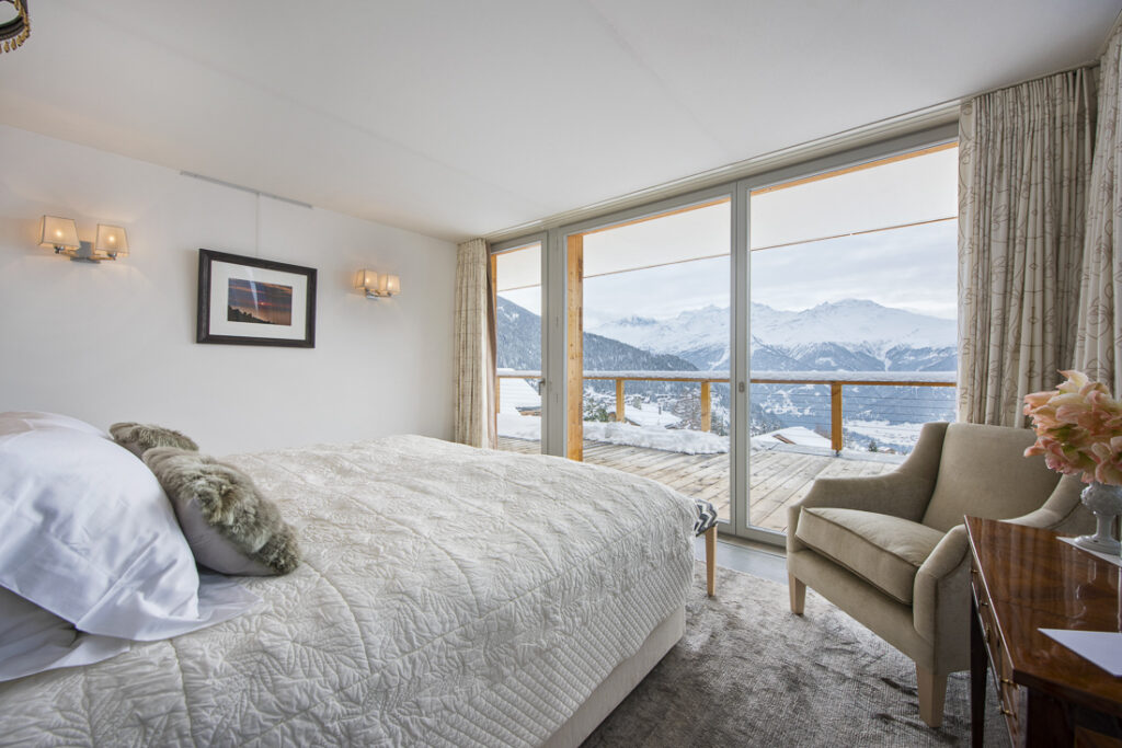 A cozy bedroom with a large bed, armchair, and bedside table with flowers, featuring floor-to-ceiling windows and a balcony overlooking snow-covered mountains and a scenic winter landscape. David Pearson Travel