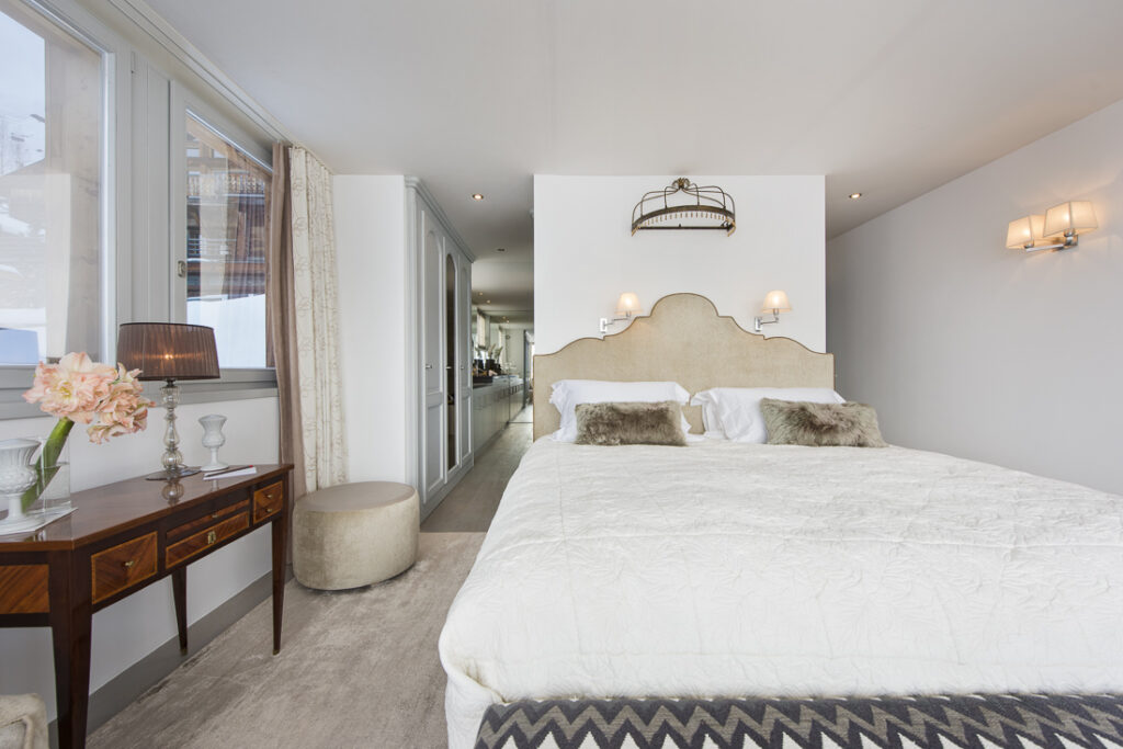 A modern, cozy bedroom with a large bed, two fur pillows, and a beige headboard. A wooden desk with flowers and a lamp sits by a window. Wall lamps provide soft lighting, and the décor is light and neutral. David Pearson Travel