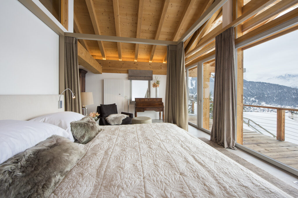 A cozy bedroom with a large bed, fur pillows, and wooden accents; floor-to-ceiling windows offer a snowy mountain view and access to a wooden balcony. Natural light fills the space, highlighting the modern rustic design. David Pearson Travel
