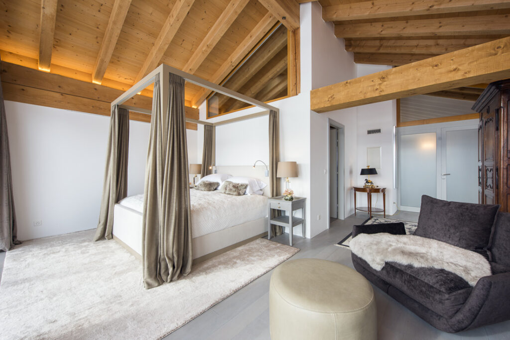 Modern bedroom with exposed wooden beams, a four-poster bed with beige curtains, white bedding, two bedside tables with lamps, a cozy gray armchair with a fur throw, and a round ottoman on a light rug. David Pearson Travel