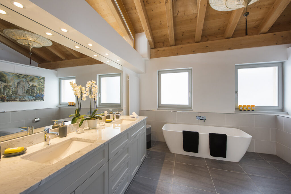 Modern bathroom with a wooden ceiling, double sink vanity with a large mirror, potted orchid, freestanding bathtub, two windows, and gray tile flooring. Towels and toiletries are neatly arranged. David Pearson Travel