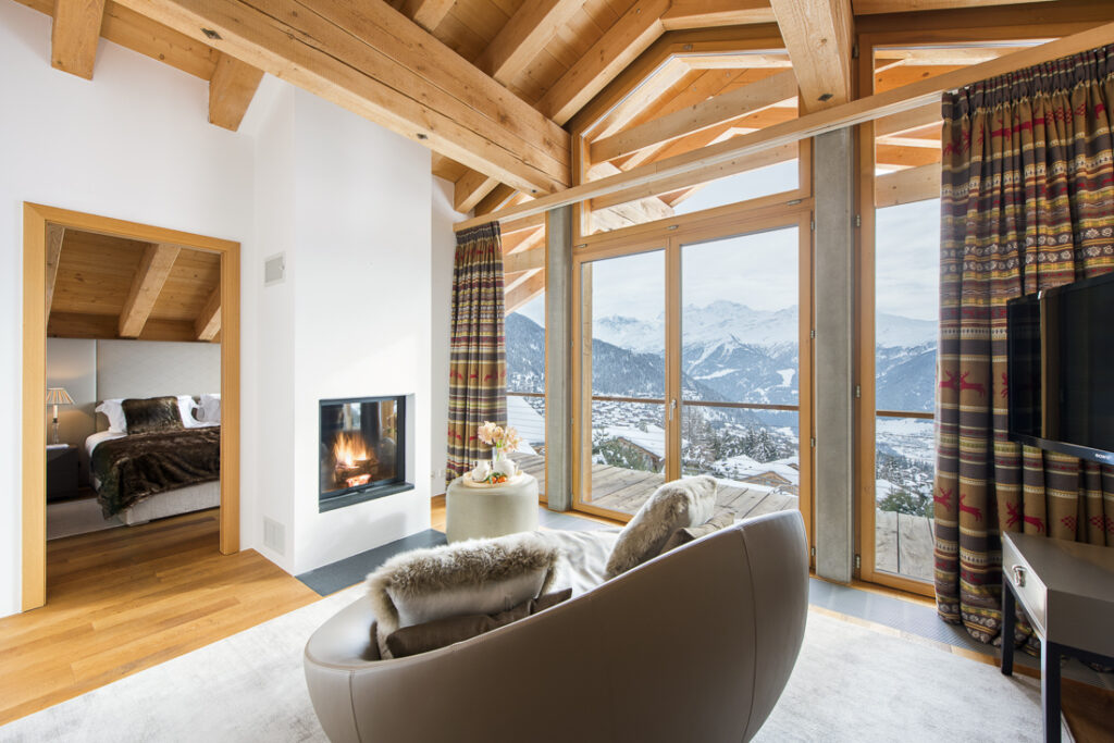 Cozy living room with a curved sofa, fur throw, and fireplace, large windows showing snowy mountain views, wooden ceiling beams, and a bedroom visible through an open doorway. David Pearson Travel