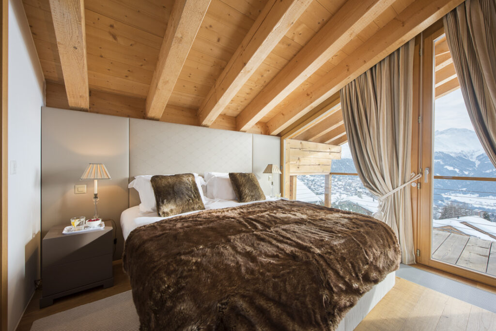 Cozy bedroom with a vaulted wooden ceiling, a large bed covered with a plush brown fur blanket, bedside tables with lamps, and floor-to-ceiling windows opening to a mountain view and wooden balcony. David Pearson Travel
