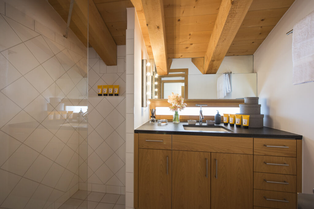 Modern bathroom with wood cabinets, a black countertop, and a sink, under a wooden ceiling with exposed beams. Shower area on the left; toiletries, towels, and flowers arranged on the right. Large mirrors reflect the space. David Pearson Travel