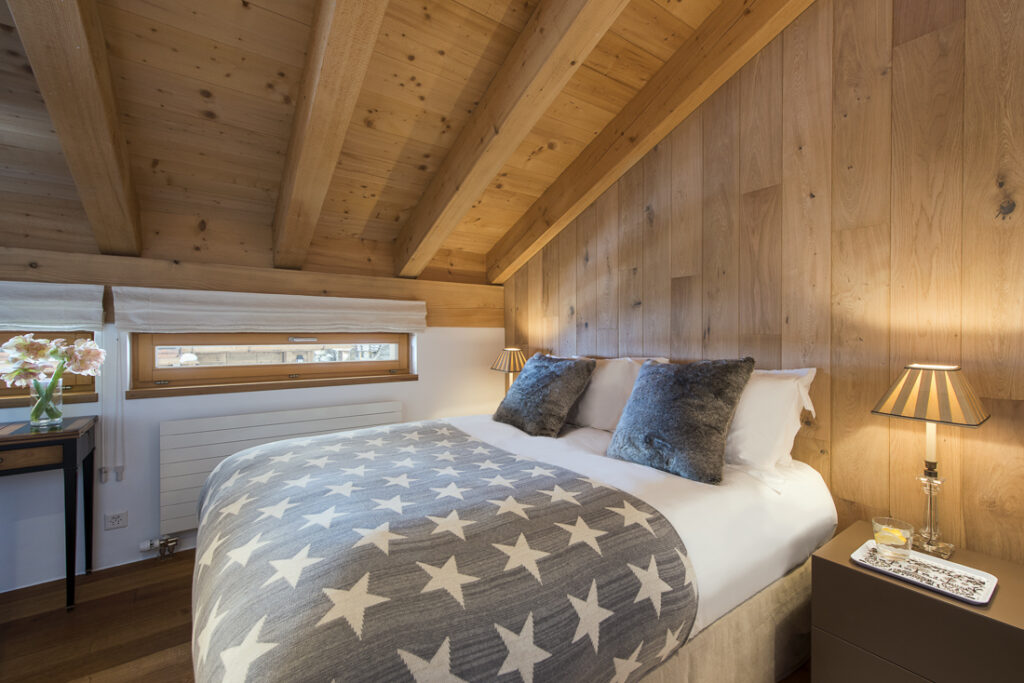 Cozy bedroom with a wooden ceiling and walls, a large bed with a star-patterned blanket, two gray pillows, bedside tables with lamps, and a window with roman shades letting in natural light. David Pearson Travel