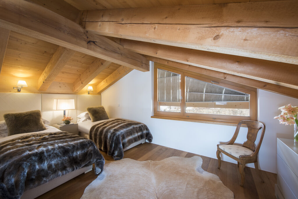 Cozy attic bedroom with exposed wooden beams, two twin beds with fur throws, a wooden chair, a window, and a cowhide rug on a wooden floor. Soft lighting and flowers add a warm, inviting feel. David Pearson Travel