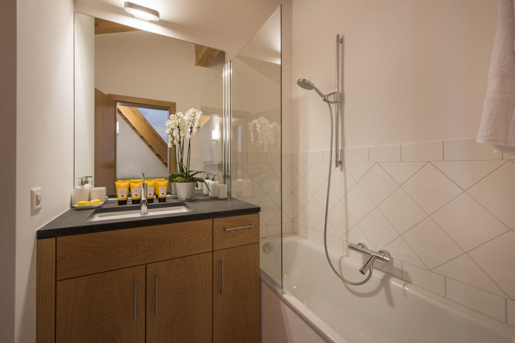 Modern bathroom with a wooden vanity, sink, and toiletries beside a vase of white orchids. A glass divider separates the sink from the bathtub with a showerhead. Rectangular mirror reflects exposed wooden beam. David Pearson Travel