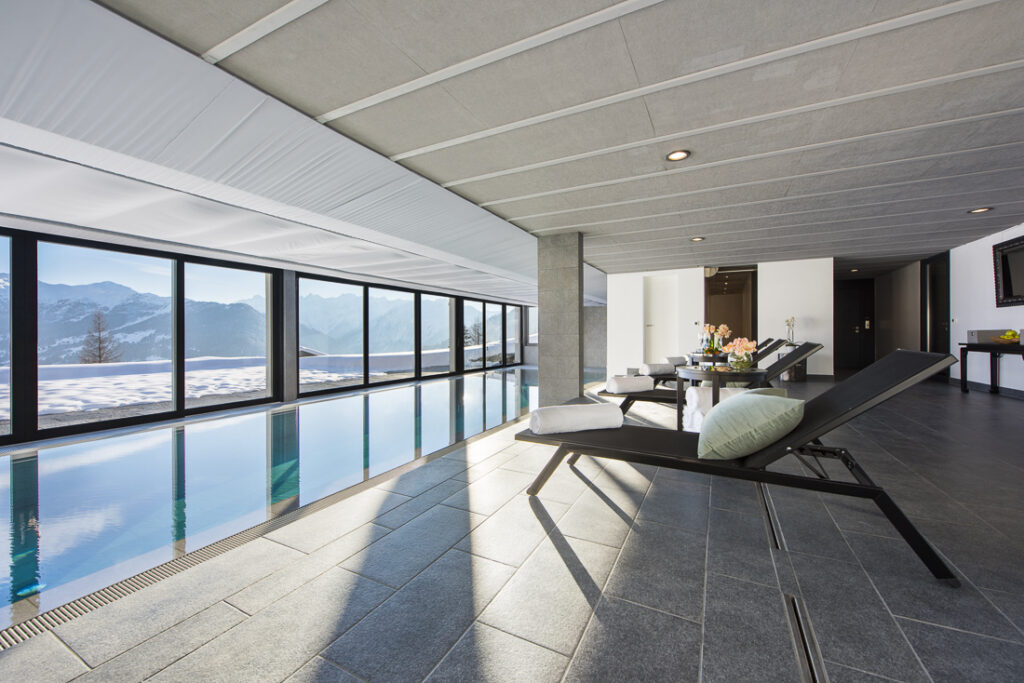 Modern indoor pool area with lounge chairs, large windows offering views of snowy mountains, and natural light filling the space. A table with flowers and drinks sits beside the pool on gray tiled flooring. David Pearson Travel