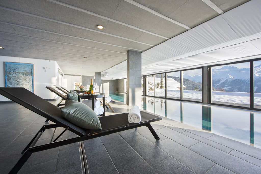 Modern indoor pool area with lounge chairs, a small table set with drinks, and large windows offering a scenic view of snowy mountains and blue sky. Rolled towels rest on one of the loungers. David Pearson Travel
