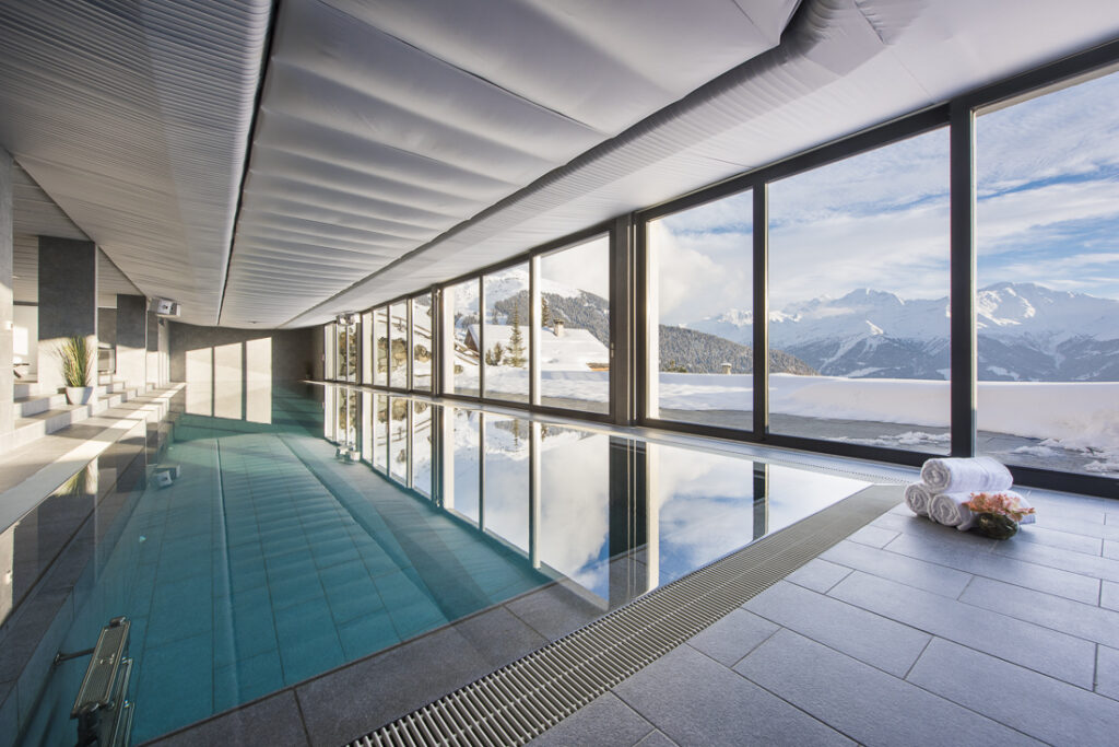 An indoor pool with large floor-to-ceiling windows overlooking snowy mountains. Rolled towels and flowers are on the tiled floor beside the pool, and sunlight streams in, creating reflections on the water. David Pearson Travel
