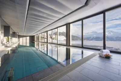 An indoor pool with large floor-to-ceiling windows overlooking snowy mountains. Rolled towels and flowers are on the tiled floor beside the pool, and sunlight streams in, creating reflections on the water. David Pearson Travel