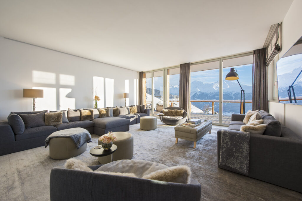 Spacious, modern living room with large windows offering mountain views. Features grey sofas with fur throws, round coffee tables, and neutral decor. Sunlight brightens the airy space. David Pearson Travel