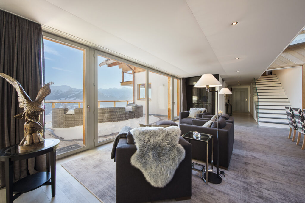 A modern, bright living room with large windows showcasing a snowy mountain view, dark armchairs with fur throws, a sculpture of an eagle, and a staircase leading to an upper floor. David Pearson Travel