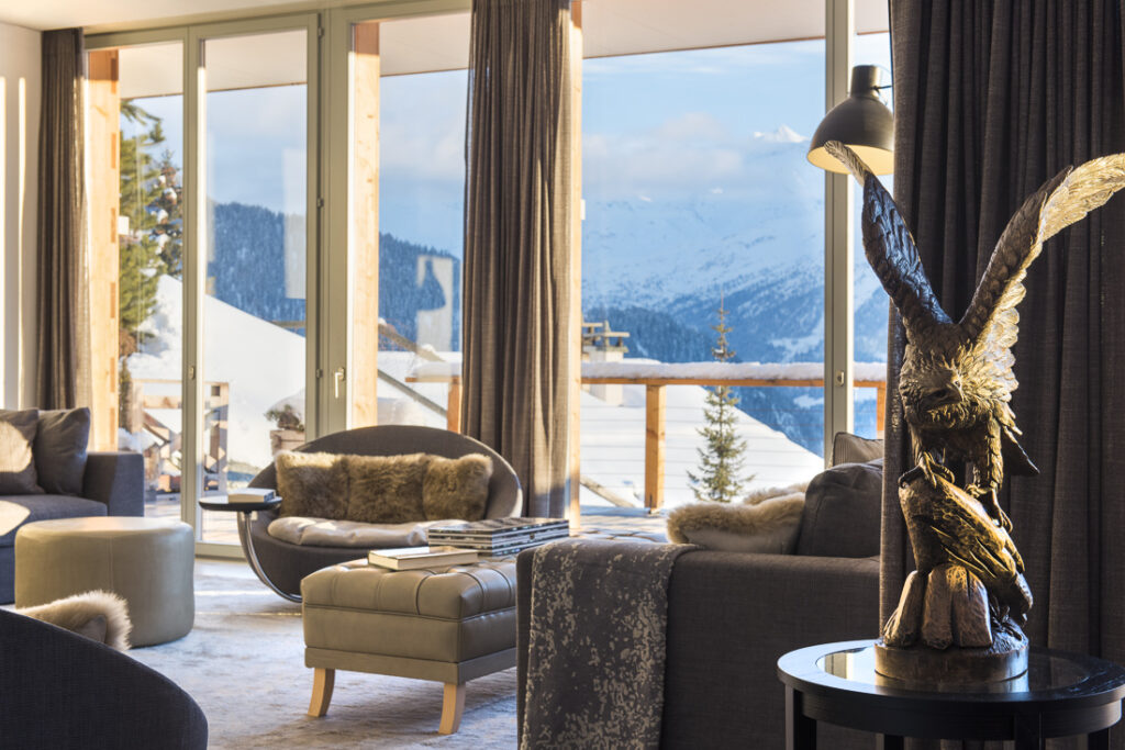 Modern, cozy living room with large windows offering a snowy mountain view. Plush chairs, fur throws, and a table with an eagle sculpture create a warm, inviting atmosphere with natural light flooding the space. David Pearson Travel