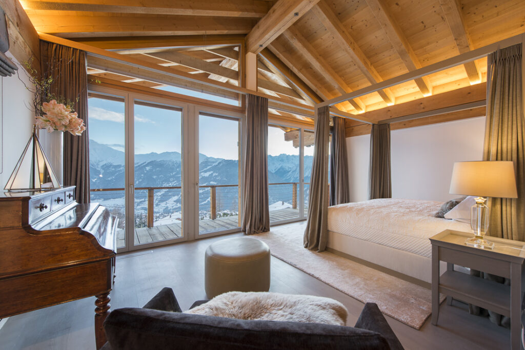 A cozy bedroom with a wooden ceiling, large windows showing snowy mountains, a bed with white linens, a side table with a lamp, a plush chair, an ottoman, and an upright piano with a vase of flowers. David Pearson Travel
