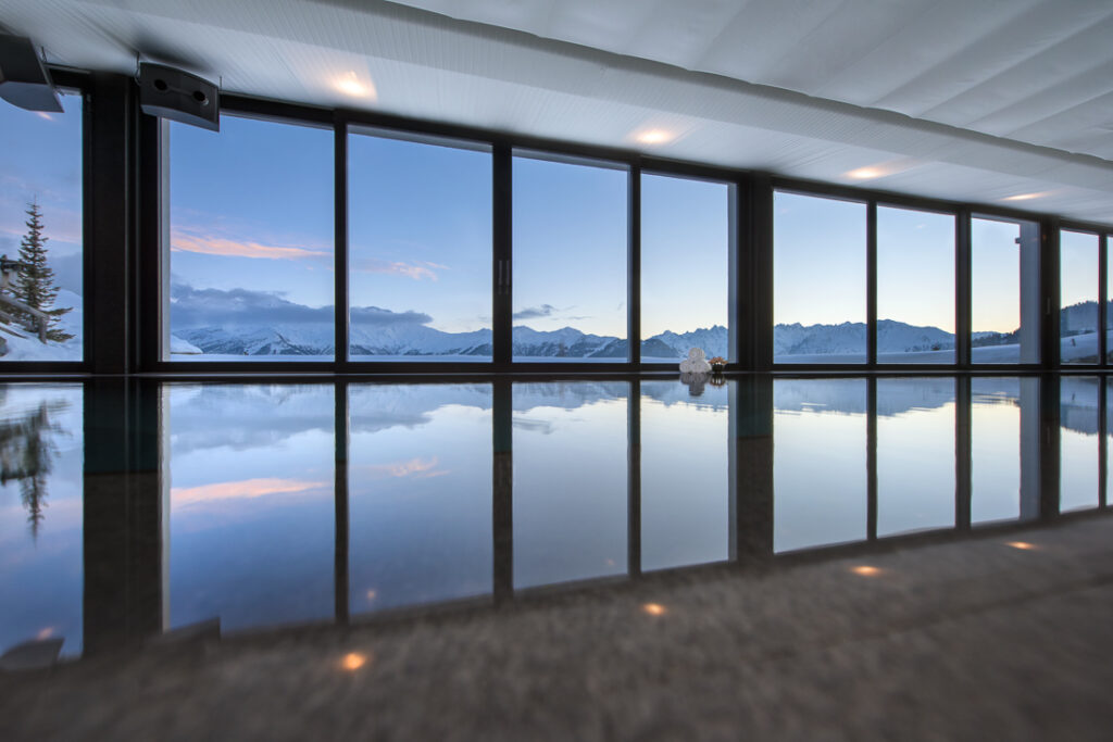 Indoor swimming pool with large floor-to-ceiling windows showing a scenic view of snow-capped mountains, blue sky, and clouds reflected in the still pool water. David Pearson Travel