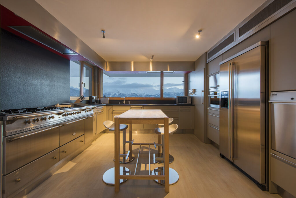 Modern kitchen with stainless steel appliances, a central wooden island with chairs, large windows showing mountain views, and light wood flooring. The space is well-lit with ceiling lights. David Pearson Travel