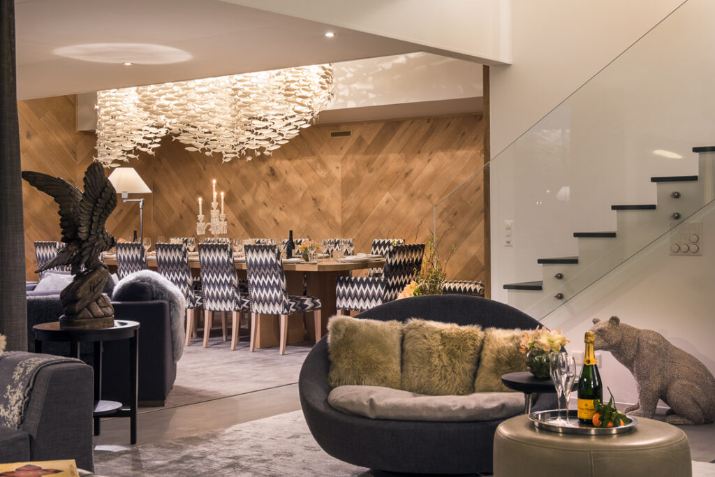 Modern living and dining area with fur-accented chairs, a round coffee table with champagne and flowers, zigzag-patterned dining chairs, wooden walls, and a striking hanging sculpture above the dining table. David Pearson Travel