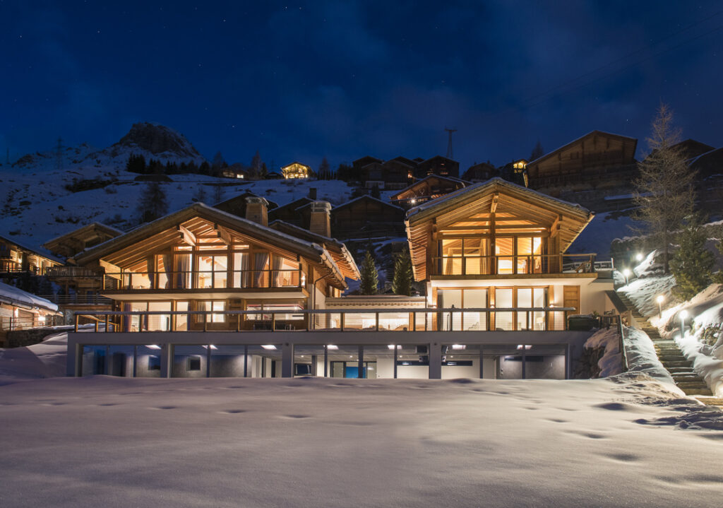 A pair of modern alpine chalets with large windows glow warmly at night, surrounded by snow and mountains, with smaller cabins and pine trees in the background. David Pearson Travel